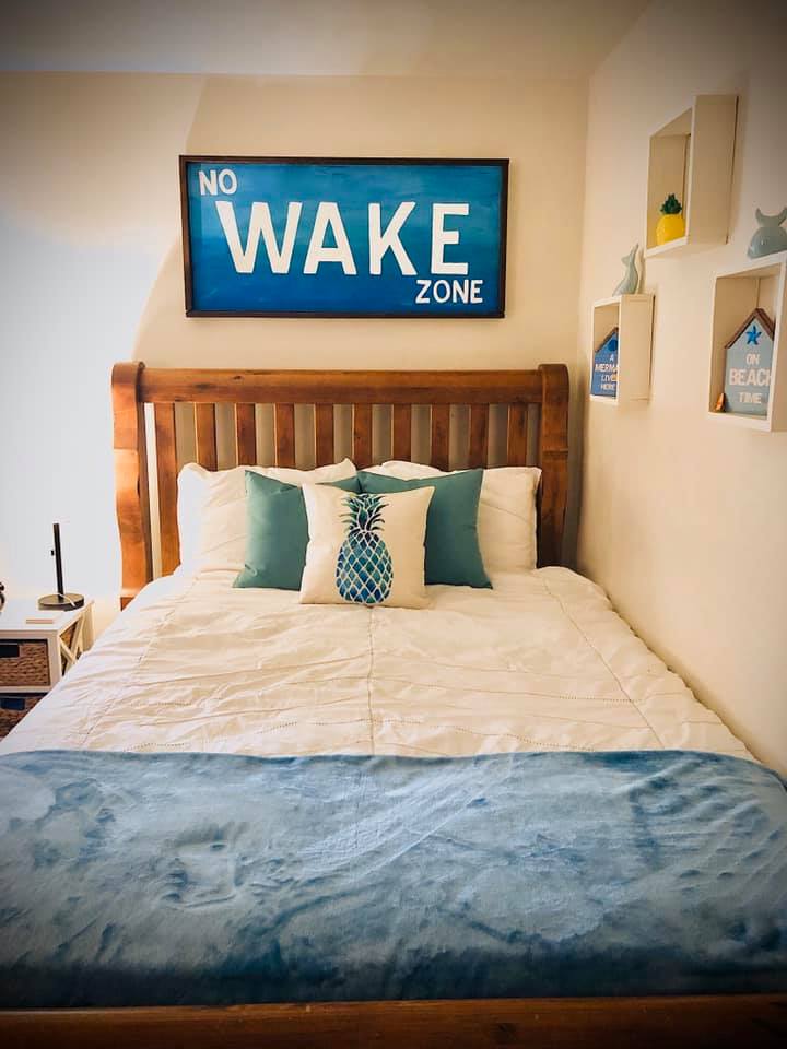 Coastal Themed Bedroom featuring white clean bedding on a natural dark wood bed frame and topped with blue throw pillows and a blanket as well as a no wake zone handmade headboard sign