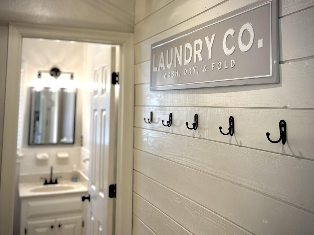 Laundry Room Design featuring shiplap wall and metal laundry co. farmhouse style sign with galvanized wall hooks. Laundry room leads to bathroom