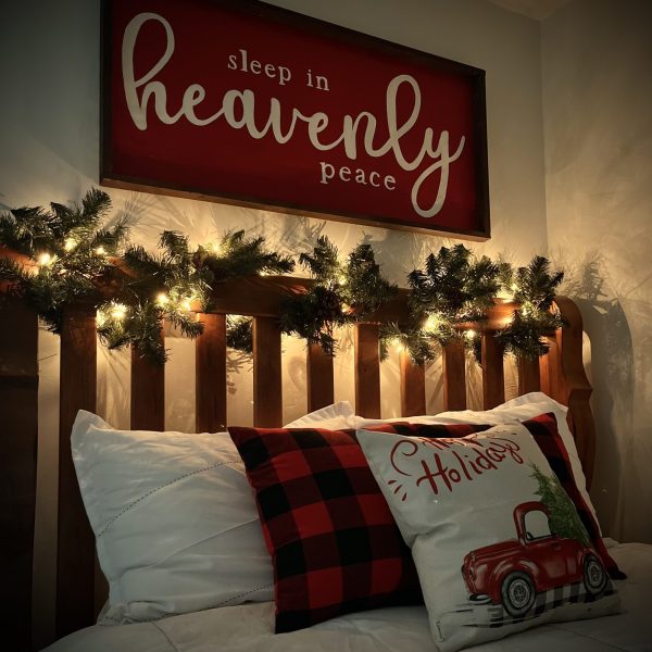 Delicate and Festive Bed Vignette for Christmas featuring a warm ambient garland woven into the headboard, a sign above the bed reading, sleep in heavenly peace, and red and black buffalo plaid throw pillows.